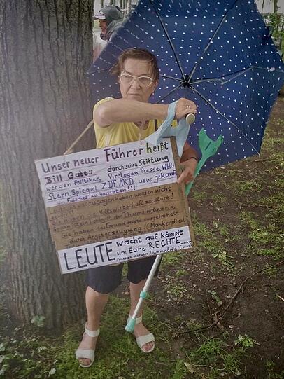 Mit Schirm, Schild und Wut auf Bill Gates: Diese Demonstrantin sucht unter einem Baum Schutz vor dem schlechten Wetter.