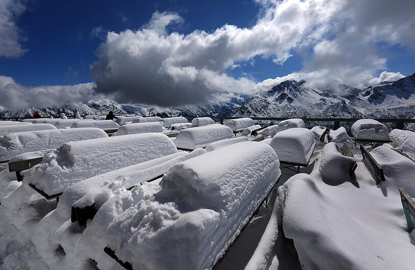 Bayern, Oberstdorf: Ausflügler stehen auf der 1780 Meter hoch gelegenen Mittelstation am Fellhorn zwischen schneebedeckten Bänken und Tischen vor dem Panorama der Alpen.