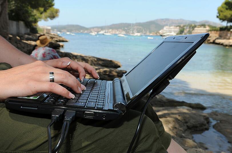 Eine Frau arbeitet am Strand von Santa Poca auf Mallorca mit ihrem Laptop am Wasser. Foto: Patrick Seeger