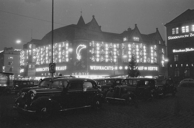 1953: Der fr&uuml;here Hertie am Hauptbahnhof unter Weihnachts-Vollbeleuchtung. Im Vordergrund vor dem Bahnhof wird geparkt.