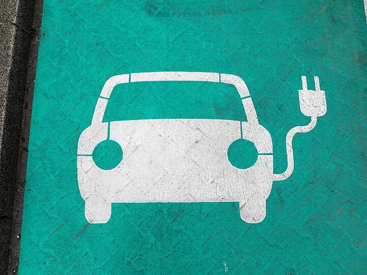 Ein stilisiertes Auto auf grünem Grund markiert einen Parkplatz mit Ladesäule für Elektrofahrzeuge. Europa und Deutschland hinken bei der Fertigung von Batteriezellen für E-Autos weit hinterher.