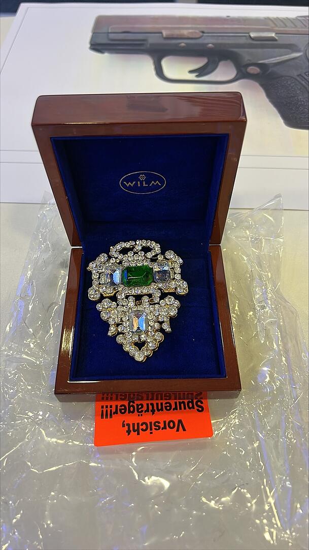 Eine unbekannte abe filigrane Brosche, dahinter nochmal das Foto der sichergestellten Waffe.