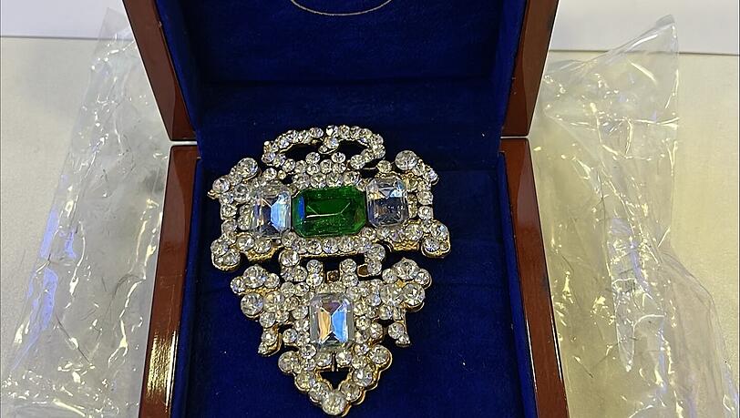 Eine unbekannte abe filigrane Brosche, dahinter nochmal das Foto der sichergestellten Waffe.