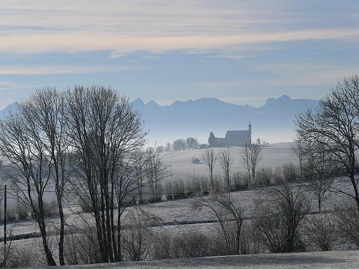 Von Frost umgeben steht die Wallfahrtskirche Sankt Alban vor dem Panorama der Alpen im Sonnenschein.