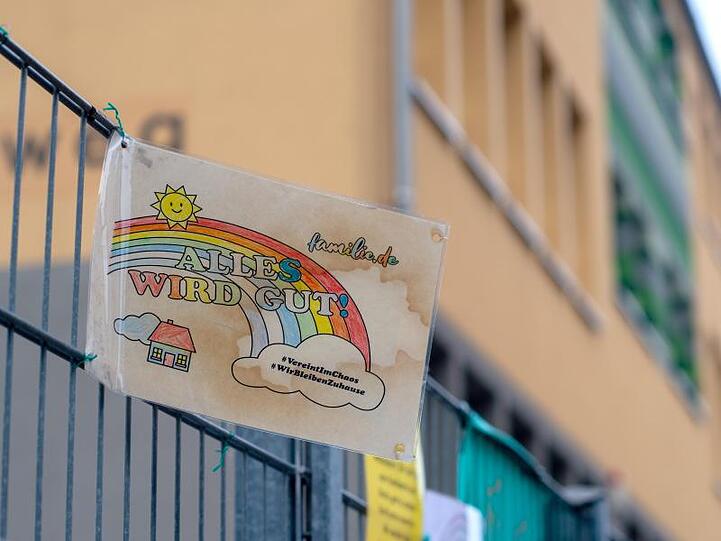 Ein Schild mit den Aufschriften "Alles wird gut!" und "#VereintImChaos" sowie "#WirBleibenZuhause" hängt am Zaun einer Grundschule.