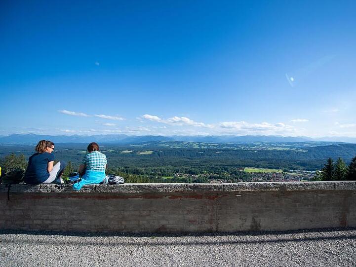 Zwei junge Frauen unterhalten sich vor dem Alpenpanorama, während sie auf einer Mauer sitzen. Foto: Lino Mirgeler/dpa/Archivbild
