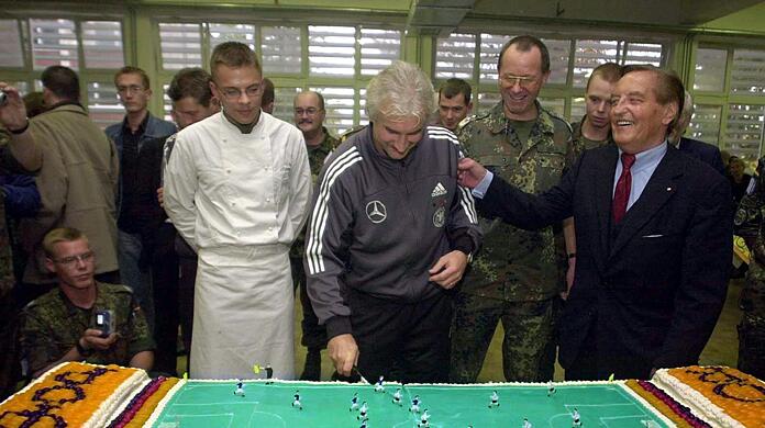 Rudi V&ouml;ller (M) schneidet beim Truppenbesuch in Sarajevo eine Torte in Form eines Fu&szlig;allplatzes an.