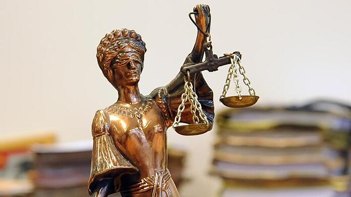 Eine goldfarbene Justitia-Figur steht vor Aktenbergen, die sich auf einem Tisch stapeln.