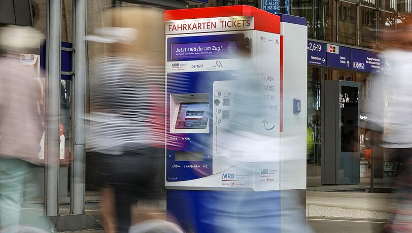 Passagiere gehen an einem Bahnhof an Fahrkartenautomaten vorbei.