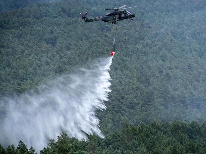 Ein Hubschrauber der Bundeswehr vom Typ NH90 mit einem Löschwasser-Behälter wirft über einer Brandstelle Löschwasser ab.