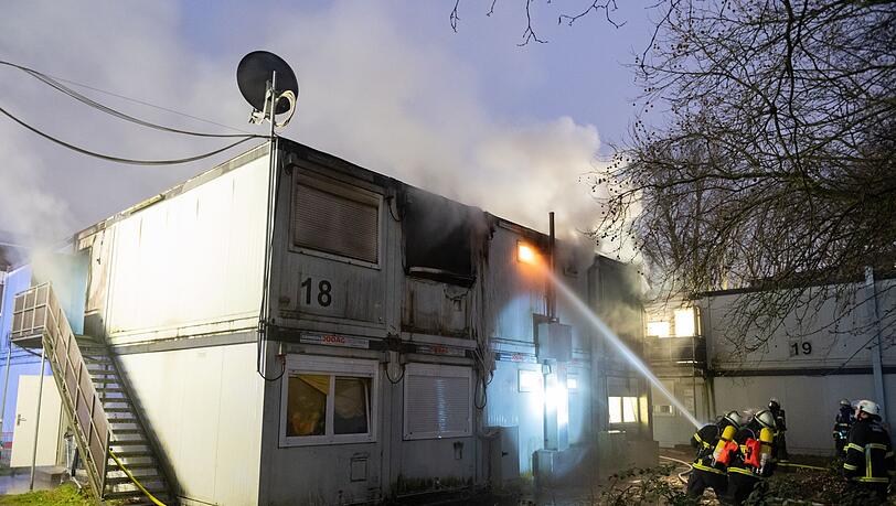 Feuerwehrleute stehen bei Löscharbeiten vor den verqualmten und teils brennenden Container-Einheiten einer Flüchtlingsunterkunft in Hamburg-Winterhude.