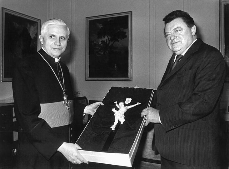 Später wurde Joseph Kardinal Ratzinger (l). als Präfekt der Glaubenskongregation, der höchsten und zentralen Instanz für die Interpretation und Verteidigung der katholischen Glaubenslehre, in den Vatikan abberufen.Der bayerische Ministerpräsident Franz Josef Strauß (r) überreicht am 12. Februar 1982 als Abschiedsgeschenk ein Kruzifix aus Porzellan an den bisherigen Erzbischof von München und Freising.