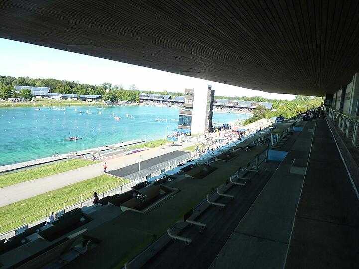 Blick von der Tribüne der Olympia-Regattaanlage auf das Becken und die Bootshallen.