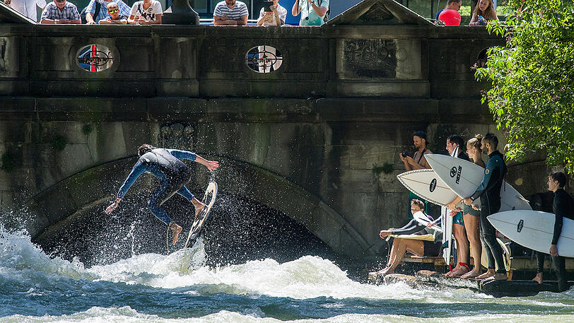 Hobbysurfer reiten die Welle auf dem Eisbach. Von der Brücke aus schauen die Touristen zu.