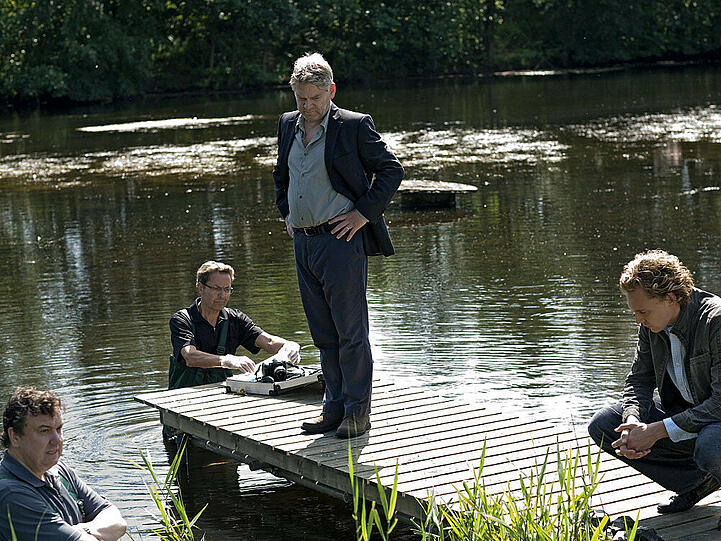 Kommissar Wallander (Kenneth Branagh, 2. v. re.) und seine Kollegen Martinsson (Tom Hiddelston, re.) und Nyberg (Richard McCabe, li.) untersuchen einen Tatort, an dem ein Mann auf grausame Weise ertränkt wurde