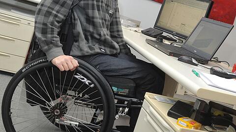 Ein Mann sitzt in einem Rollstuhl an seinem Arbeitsplatz am Schreibtisch.