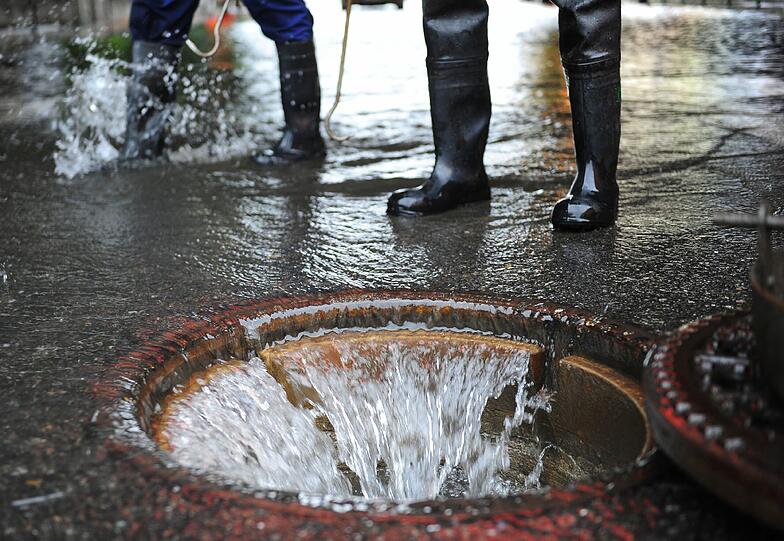 Gurgelnd schluckt der Gully die Wassermassen &ndash; dahinter stehen Feuerwehrler in Gummistiefeln.