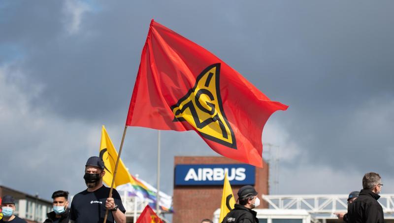 Beschäftigte eines Airbus-Werkes stehen vor den Toren des Werksgeländes und schwenken Fahnen der IG Metall.