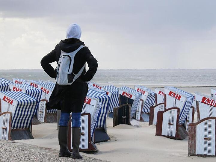 Grau und windig auf Norderney: Eine Frau steht warm angezogen auf der Strandpromenade und schaut über die verwaisten Strandkörbe hinweg auf die Nordsee.