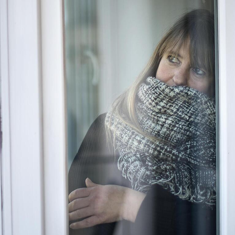 Eine Frau blickt warm angezogen aus ihrem Wohnungsfenster. In M&uuml;nchen wird es ab Donnerstag kalt.
