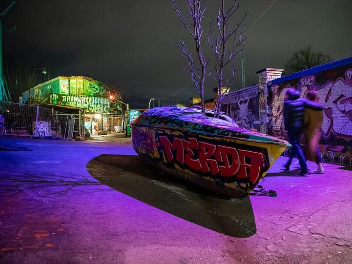 Personen gehen an einem ausrangierten Boot vor dem Eingang des Nachtclubs "Bahnwärter Thiel". Foto: Lino Mirgeler/dpa