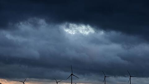 Windräder stehen bei Aitrang im Allgäu unter dichten Regenwolken hinter einem Wald.