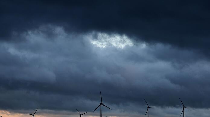 Windräder stehen bei Aitrang im Allgäu unter dichten Regenwolken hinter einem Wald.