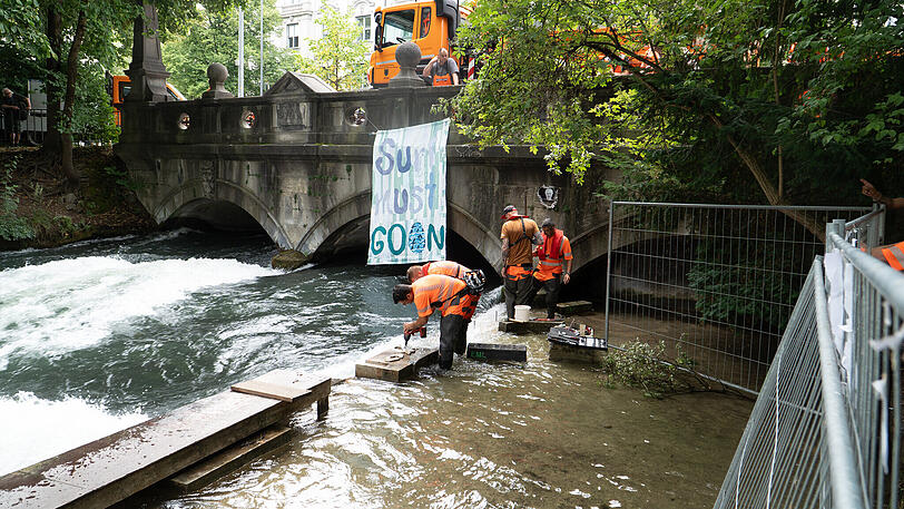 Betonkl&ouml;tze und Absperrungen werden am Eisbach entfernt. Im Hintergrund ist ein Banner mit der Aufschrift &laquo;Surf must go on&raquo; an der Eisbachbr&uuml;cke an der Eisbachwelle befestigt. Seit dem Tod einer Surferin im M&uuml;nchner Eisbach war die Welle gesperrt. Die Ermittlungen sind beendet - und jetzt auch die Wartezeit f&uuml;r die Wellenreiter. Doch f&uuml;r sie gibt es neue Regeln.