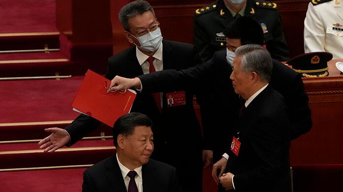 Chinas Ex-Präsident Hu Jintao (r) wird von seinem Platz weg begleitet. Links sitzt Xi Jinping und guckt zu.
