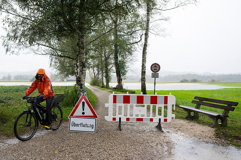 Felden Am Chiemsee: Ein Radfahrer fährt bei Felden am Chiemsee an einem Schild mit der Aufschrift "Überflutung" vorbei. Der Hauptteil der fürs Wochenende angekündigten Niederschläge in Bayern ist nach Erkenntnis der Hochwassernachrichtenzentrale bereits gefallen. Der Dauerregen der vergangenen Stunden habe im Südosten Bayerns die Wasserstände vom Isar-Einzugsgebiet bis zum Bayerischen Wald ansteigen lassen, an einigen Orten gebe es kleinere Ausuferungen.