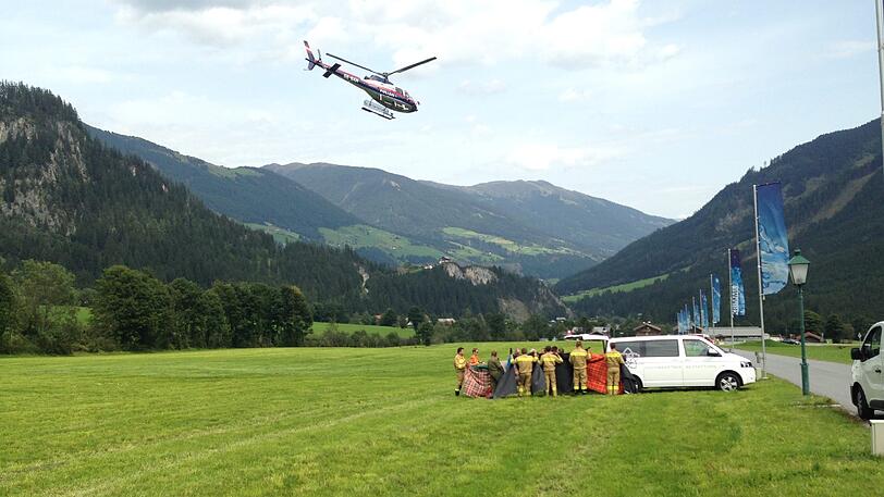 Ein Rettungshelikopter fliegt über ein Feld in Krimml, während Einsatzkräfte an einem Auto stehen.