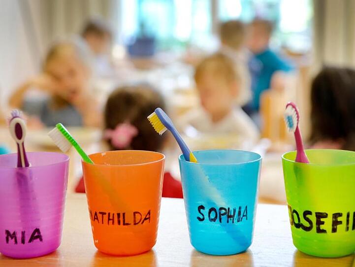 Zahnbürsten und Zahnputzbecher mit den Vornamen der Kinder stehen in einer Kindertagesstätte auf einem Tisch.