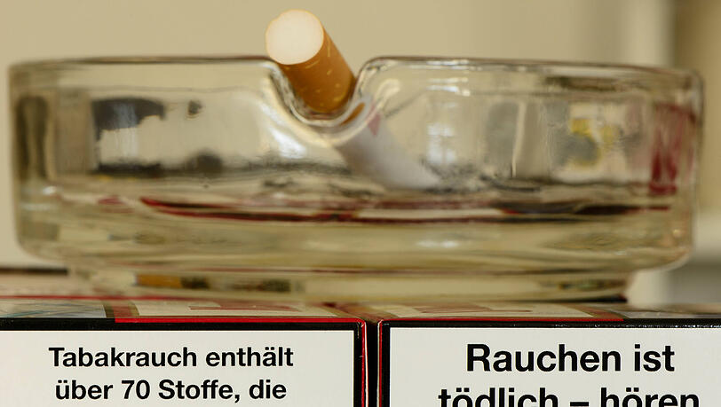 Auf zwei Zigarettenpackungen mit Gesundheitswarnung steht ein Aschenbecher. Auf zwei Zigarettenpackungen mit Gesundheitswarnung steht ein Aschenbecher.