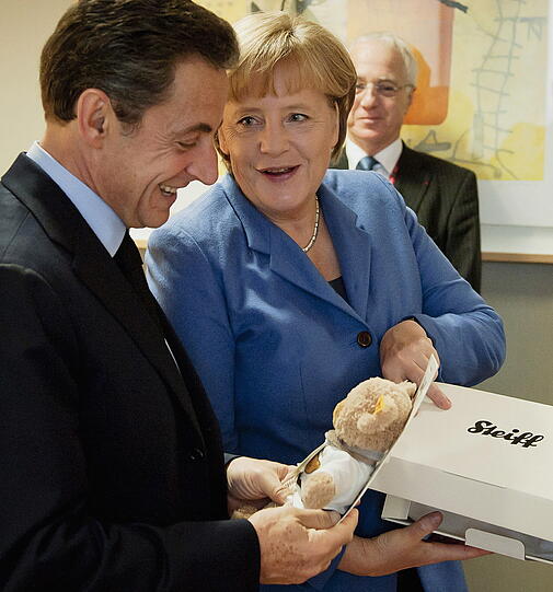 Baby-Geschenk: Angela Merkel überreicht Sarkozy hinter den Kulissen einen Teddybären für die kleine Giulia.