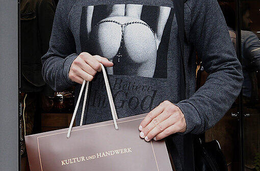 "Believe in God" steht auf "Stoppels" T-Shirt. Unfreiwillige Komik: Auf der Meindl-Einkaufstasche ist der Slogan: "Kultur und Handwerk" aufgedruckt. Darüber das T-Shirt mit dem fast nackten Damen-Hintern.