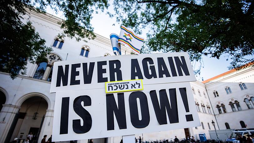 Ein Schild mit der Aufschrift „Never again is now!“ ist bei Teilnehmern einer Mahnwache gegen ein geplantes Pro-Palästina-Protestcamp vor der Ludwig-Maximilians-Universität (LMU) zu sehen.