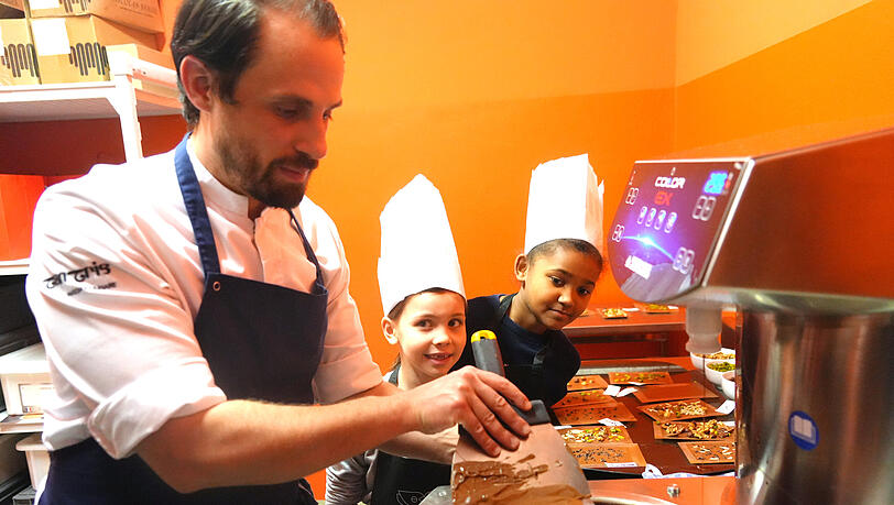Wer an der Schokoladen- Quelle steht, bekommt die ganze Aufmerksamkeit. Chef-Patissier Maxime Rebmann zeigt den Schülern, wie aus der flüssigen Schokolade eine knackige Tafel wird. Das dürfen die Kinder natürlich auch selbst ausprobieren und ihre Tafeln anschließend mit Pistazien, Mandeln und mehr bestücken.
