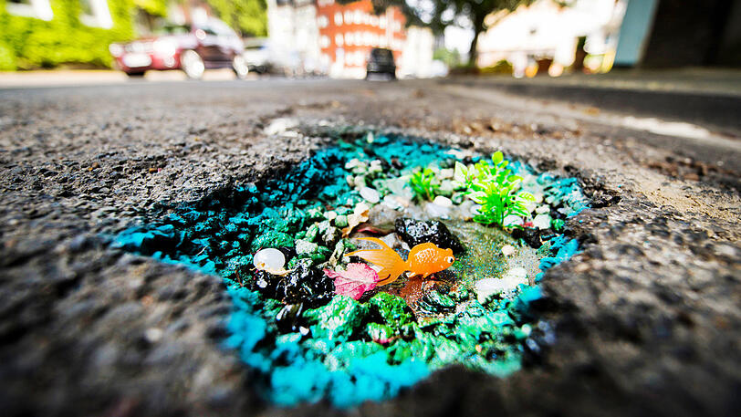 27.07.2018: In einem Schlagloch einer Straße liegt ein Goldfisch aus Plastik, umgeben von kleinen Pflanzen und glitzerndem Harz. Eine Street-Art Künstlerin hat das Schlagloch am Straßenrand in ein kleines Kunstwerk verwandelt.
