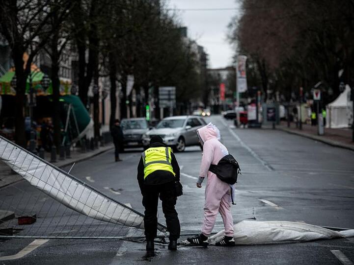 Ein Ordner und ein verkleideter Narr stehen in Düsseldorf an einem Bauzaun, der durch den Sturm umgeweht worden ist.