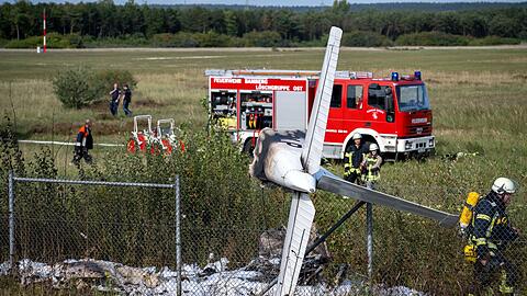 Trümmerteile eines Flugzeugs hängen in einem Zaun, während Einsatzkräfte der Feuerwehr an der Absturzstelle stehen.
