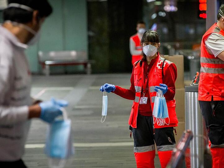 Freiwillige Mitarbeiter des Roten Kreuzes verteilen vor dem Bahnhof Barcelona-Catalunya Mundschutzmasken an Passagiere.