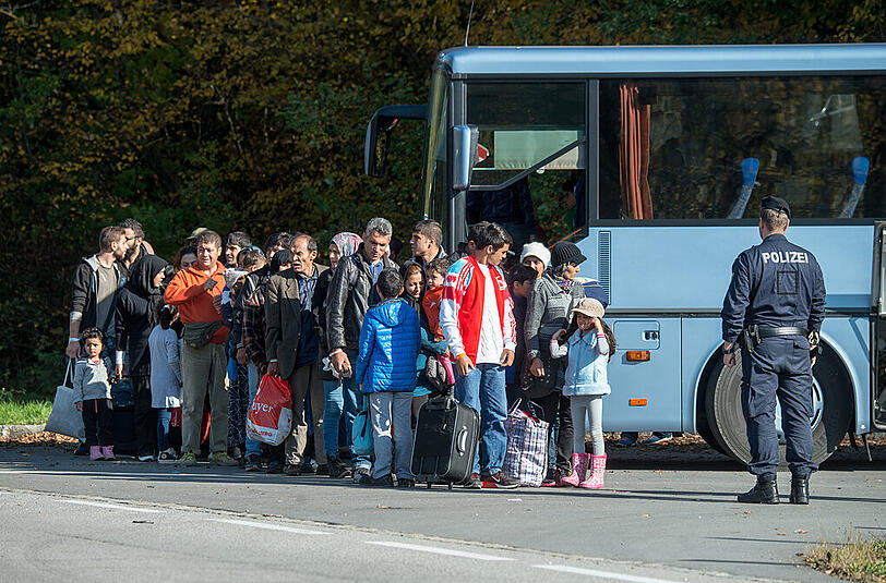 Flüchtlinge warten neben einem Bus. Ein österreichischer Beamter sieht zu.