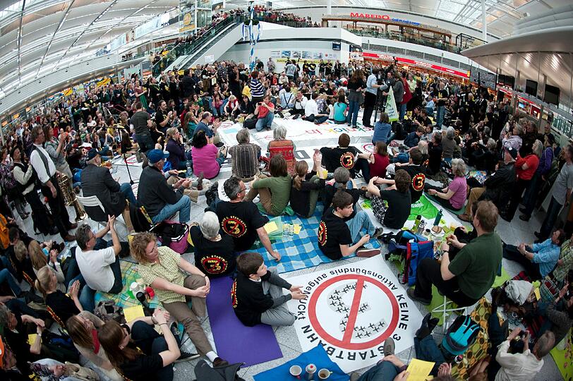 Demonstranten sitzen am Samstag (24.03.2012) am Terminal 2 des Münchner Flughafens auf Picknickdecken. Der Bund Naturschutz und das Aktionsbündnis AufgeMUCkt haben zum Protest-Picknick aufgerufen, um gegen den Bau einer dritten Startbahn am Münchner Flughafen zu demonstrieren.
