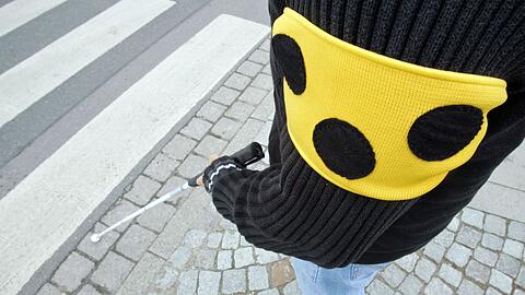 Ein blinder Mann geht mit seinem weißen Taststock und einer Armbinde mit drei Punkten über eine Straße in Hannover.