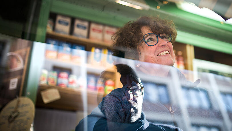 Marktfrau Marina Bröckelt lächelt in ihrem Stand auf dem Viktualienmarkt in München, während sich im Tresen ein Kunde spiegelt. Marktfrau Marina Bröckelt lächelt in ihrem Stand auf dem Viktualienmarkt in München, während sich im Tresen ein Kunde spiegelt.