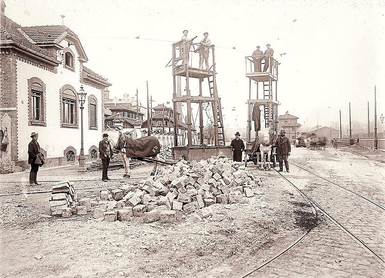 Pferde helfen beim Transport von Pflastersteinen, während Arbeiter die Tram-Oberleitung installieren.