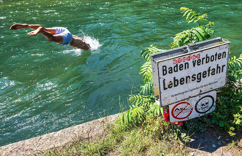 Eine Person springt in den Eisbach im Englischen Garten. Am Uferrand steht ein Schild mit der Aufschrift „Baden verboten". Eine Person springt in den Eisbach im Englischen Garten. Am Uferrand steht ein Schild mit der Aufschrift „Baden verboten".