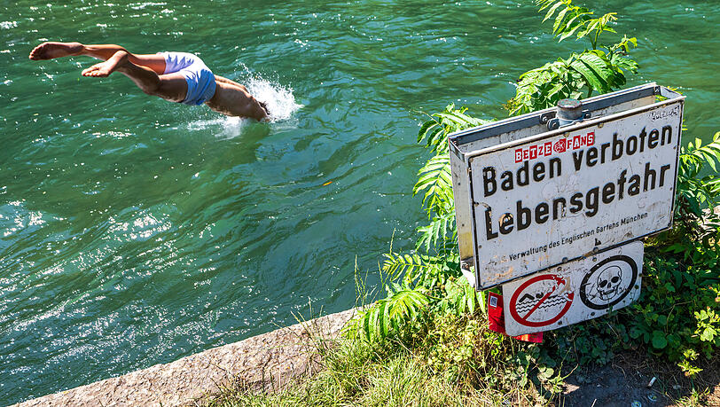 Eine Person springt in den Eisbach im Englischen Garten. Am Uferrand steht ein Schild mit der Aufschrift „Baden verboten". Eine Person springt in den Eisbach im Englischen Garten. Am Uferrand steht ein Schild mit der Aufschrift „Baden verboten".