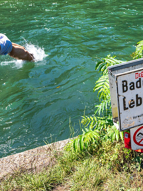 Eine Person springt in den Eisbach im Englischen Garten. Am Uferrand steht ein Schild mit der Aufschrift &bdquo;Baden verboten".