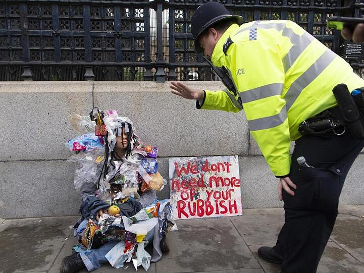 Ein Polizist spricht in London mit einer mit Müll bedeckten Klima-Aktivistin, die neben einem Plakat mit der Aufschrift "We don't need more of your rubbish" (Wir brauchen nicht mehr von eurem Müll) sitzt.